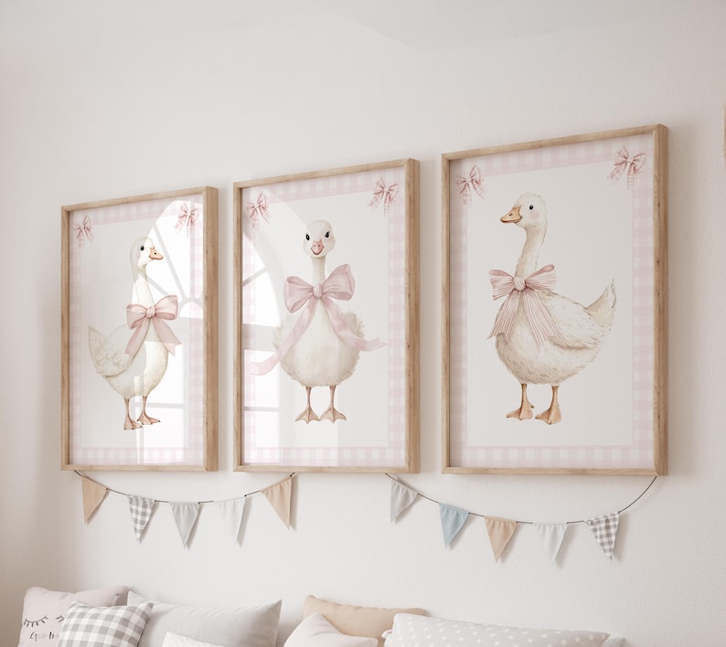 K&ouml;nnte beinhalten: Drei gerahmte Aquarelldrucke von G&auml;nsen, jede mit einer rosa Schleife. Die Drucke haben einen wei&szlig;en Hintergrund mit einem rosa Vichy-Rand und einem Holzrahmen. Unterhalb der Rahmen h&auml;ngt eine dekorative Wimpelkette.