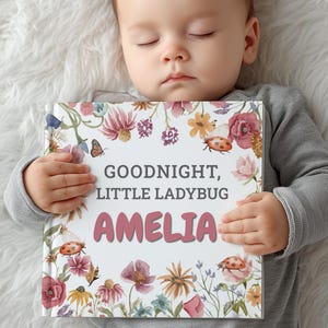 May include: A square hardcover book titled Goodnight, Little Ladybug with the name Amelia printed in large pink letters. The cover features a white background with watercolor illustrations of red ladybugs, butterflies, and various colorful flowers.