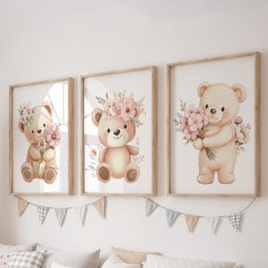 May include: Three framed watercolor prints of teddy bears. Each bear is a light brown color and holds or wears pink flowers. The frames are light brown and hang on a white wall. A garland of small flags hangs below the frames.