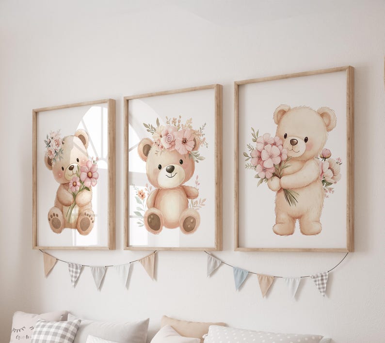 May include: Three framed watercolor prints of teddy bears. Each bear is a light brown color and holds or wears pink flowers. The frames are light brown and hang on a white wall. A garland of small flags hangs below the frames.