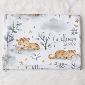 May include: A white baby blanket featuring a repeating pattern of sleeping cheetahs, clouds, stars, and foliage in muted tones. The name "William JAMES" is printed in gray script. The blanket is soft and cozy.