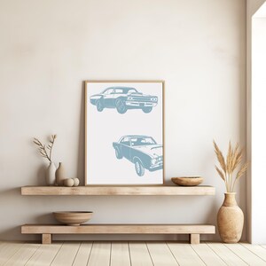 Pode incluir: Uma impress&atilde;o emoldurada com um design minimalista que apresenta duas silhuetas azuis de carros cl&aacute;ssicos americanos de m&uacute;sculo. A impress&atilde;o est&aacute; pendurada em uma parede branca acima de uma prateleira de madeira.