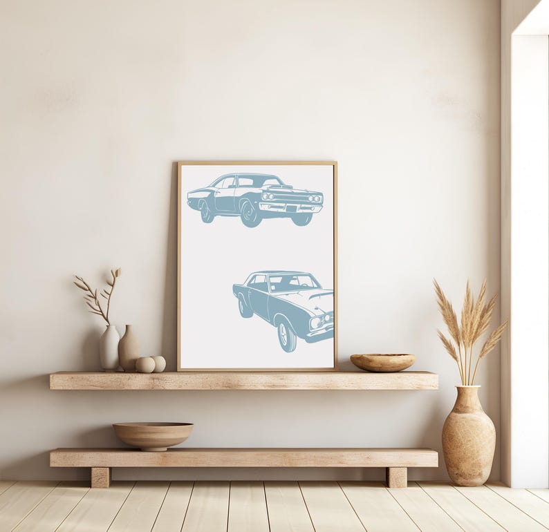 Pode incluir: Uma impress&atilde;o emoldurada com um design minimalista que apresenta duas silhuetas azuis de carros cl&aacute;ssicos americanos de m&uacute;sculo. A impress&atilde;o est&aacute; pendurada em uma parede branca acima de uma prateleira de madeira.
