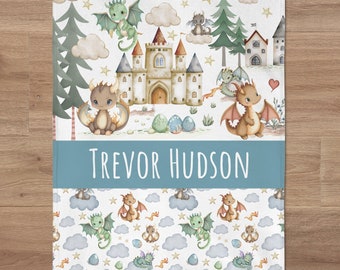 May include: A personalized baby blanket with a blue and white border. The center of the blanket features a watercolor illustration of a castle, trees, and dragons. The bottom border has the name "Trevor Hudson" in white text. The blanket has a repeating pattern of dragons, clouds, and stars.