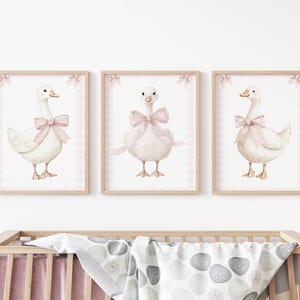 K&ouml;nnte beinhalten: Drei gerahmte Aquarelldrucke mit wei&szlig;en G&auml;nsen, die mit rosa Schleifen geschm&uuml;ckt sind. Jede Gans ist vor einem wei&szlig;en Hintergrund mit einem rosa Vichy-Rand und einem Holzrahmen platziert. Geeignet f&uuml;r ein Kinderzimmer.