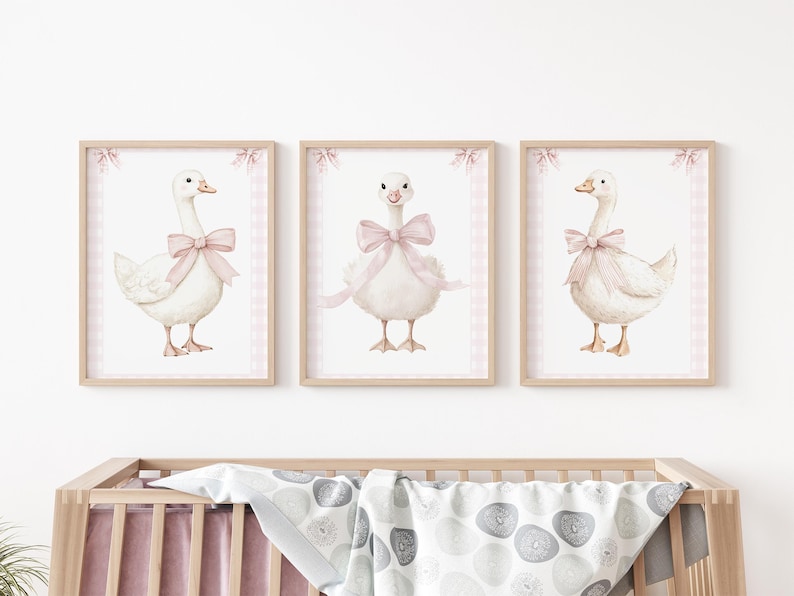 K&ouml;nnte beinhalten: Drei gerahmte Aquarelldrucke mit wei&szlig;en G&auml;nsen, die mit rosa Schleifen geschm&uuml;ckt sind. Jede Gans ist vor einem wei&szlig;en Hintergrund mit einem rosa Vichy-Rand und einem Holzrahmen platziert. Geeignet f&uuml;r ein Kinderzimmer.