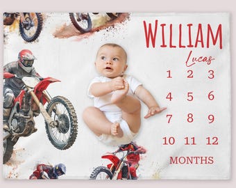 May include: White baby milestone blanket featuring watercolor illustrations of motocross bikes and riders. The blanket includes the name "William Lucas" in red script, along with numbers 1-12 and the word "MONTHS". A baby is lying on the blanket.