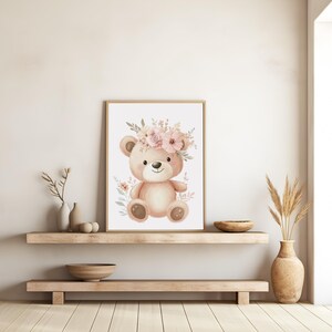 May include: A framed watercolor print of a teddy bear wearing a floral crown. The bear is light brown with a smiling face and sits against a white background. Soft pink and green floral accents surround the bear, creating a gentle, nursery-themed artwork.