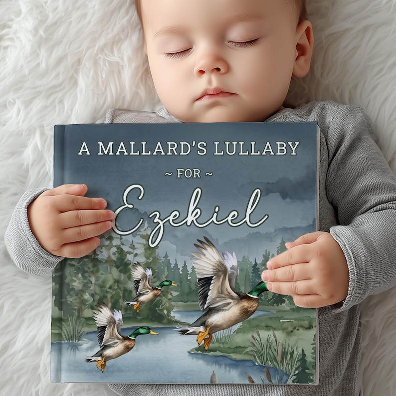 Może przedstawiać: Książka w twardej oprawie zatytułowana A Mallard's Lullaby for Ezekiel z akwarelową ilustracją trzech krzyż&oacute;wek lecących nad rzeką na tle lasu. Okładka książki ma ciemnoniebieskie niebo i biały tekst.