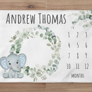 May include: A white baby milestone blanket with a green eucalyptus wreath, a gray elephant with blue polka dots, and a numbered chart for tracking a baby's growth. The text "ANDREW THOMAS" is at the top of the blanket.