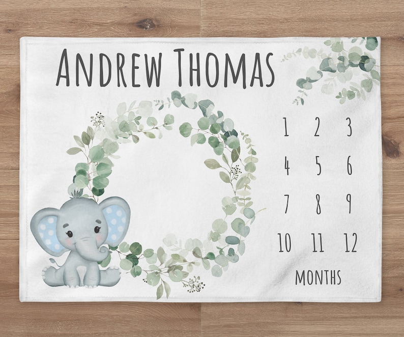 May include: A white baby milestone blanket with a green eucalyptus wreath, a gray elephant with blue polka dots, and a numbered chart for tracking a baby's growth. The text "ANDREW THOMAS" is at the top of the blanket.