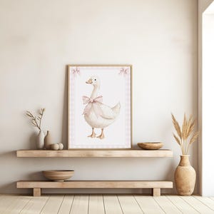 K&ouml;nnte beinhalten: Ein gerahmter Aquarelldruck mit einer wei&szlig;en Gans, die mit einer rosa Schleife geschm&uuml;ckt ist. Das Kunstwerk ist vor einem rosa Vichy-Rand platziert und in hellem Holz gerahmt. Der Druck wird auf einem Holzregal ausgestellt und verleiht der Einrichtung einen rustikalen Charme.