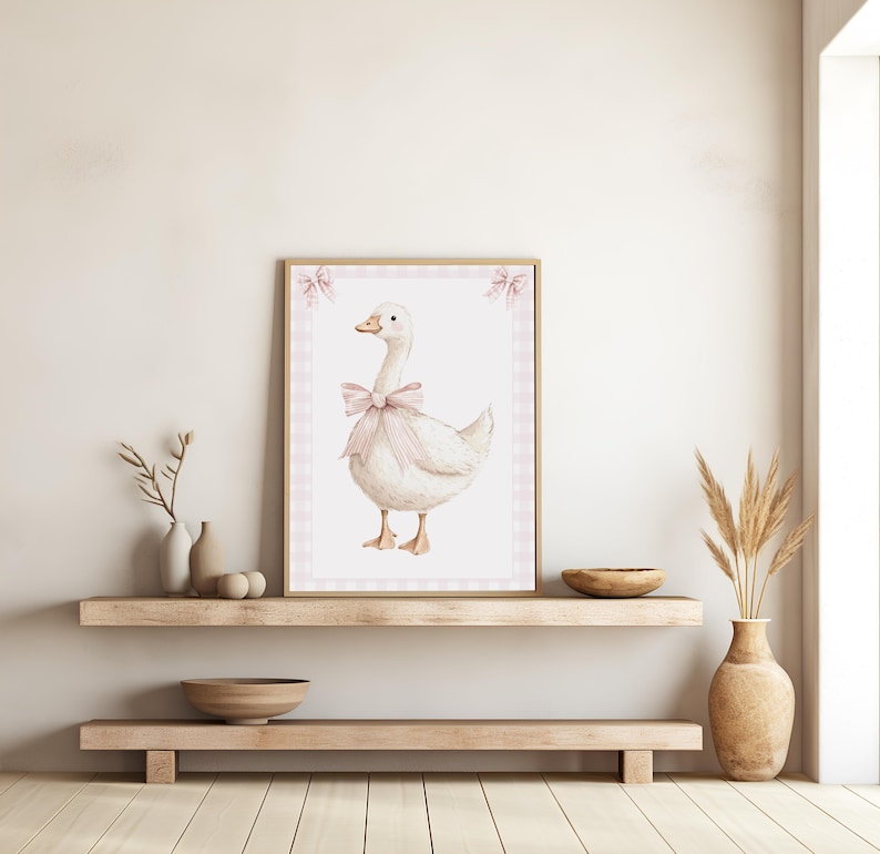 K&ouml;nnte beinhalten: Ein gerahmter Aquarelldruck mit einer wei&szlig;en Gans, die mit einer rosa Schleife geschm&uuml;ckt ist. Das Kunstwerk ist vor einem rosa Vichy-Rand platziert und in hellem Holz gerahmt. Der Druck wird auf einem Holzregal ausgestellt und verleiht der Einrichtung einen rustikalen Charme.