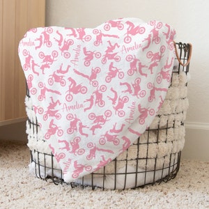 May include: Pink and white baby blanket with a pattern of pink silhouettes of people riding dirt bikes. The blanket has the name "Amelia" repeated throughout the pattern.