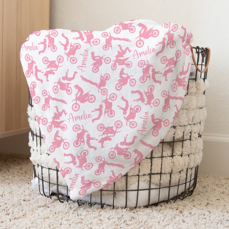 May include: Pink and white baby blanket with a pattern of pink silhouettes of people riding dirt bikes. The blanket has the name "Amelia" repeated throughout the pattern.