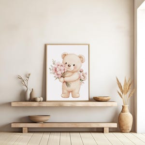 May include: A framed watercolor print featuring a beige teddy bear holding a bouquet of pink flowers. The artwork has a soft, pastel color palette and is displayed on a light-colored wall above a wooden shelf.