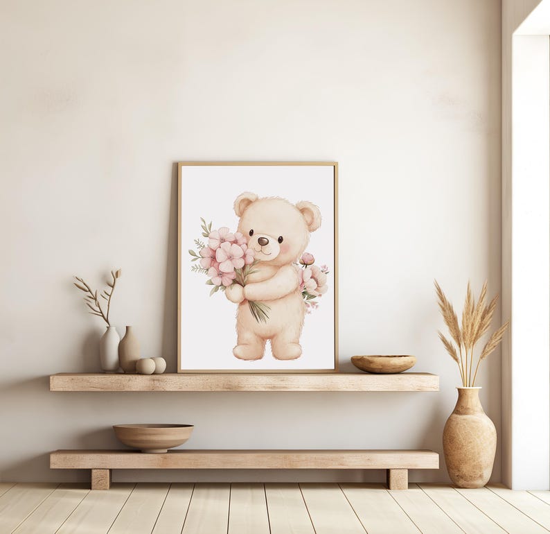 May include: A framed watercolor print featuring a beige teddy bear holding a bouquet of pink flowers. The artwork has a soft, pastel color palette and is displayed on a light-colored wall above a wooden shelf.