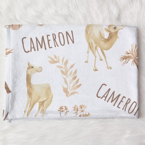 May include: A white baby blanket with a brown camel and llama pattern. The blanket has the name "CAMERON" printed on it twice.