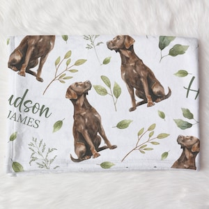 May include: White blanket featuring a repeating pattern of brown Labrador Retrievers, green leaves, and sprigs. The blanket has the name "Hudson James" in a script font. The overall design is a watercolor style.