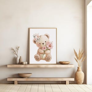 May include: A framed watercolor print of a teddy bear holding pink flowers and wearing a floral crown. The bear is light brown with dark brown accents. The artwork is displayed on a light-colored wall above a wooden shelf.