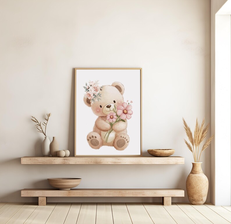 May include: A framed watercolor print of a teddy bear holding pink flowers and wearing a floral crown. The bear is light brown with dark brown accents. The artwork is displayed on a light-colored wall above a wooden shelf.