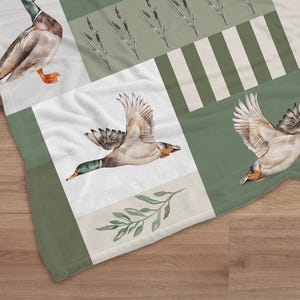 May include: A soft, square blanket featuring a watercolor design of ducks in flight and reeds. The blanket has a patchwork pattern with shades of green, white, and beige. The ducks are depicted with brown, white, and green feathers.