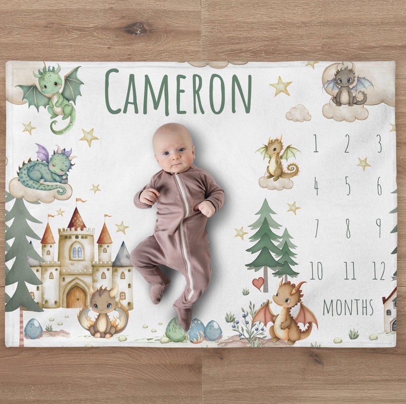 May include: A white baby milestone blanket with a green, brown, and gold castle, stars, and dragons. The blanket has a number chart for each month of the year. The name "CAMERON" is written in green letters at the top of the blanket. A baby is lying on the blanket.