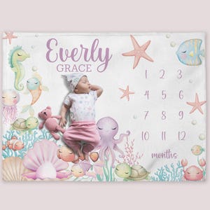 May include: A white baby blanket with a sea-themed design. It features a baby girl with a purple octopus, seahorses, starfish, seashells, and a turtle. The blanket also has a monthly growth chart with numbers 1-12. The text "Everly Grace" is printed on the blanket.