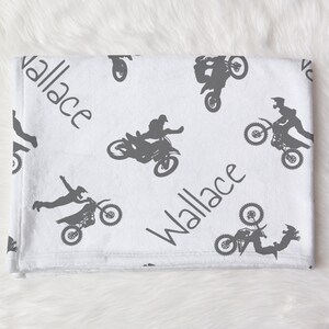 K&ouml;nnte beinhalten: Wei&szlig;e Babydecke mit grauem Motocross-Design. Die Decke zeigt sich wiederholende Bilder von Motocross-Fahrern und Motorr&auml;dern sowie den Namen "Wallace" in Handschrift.