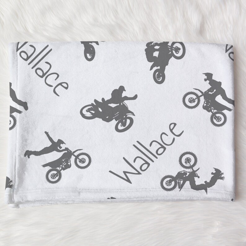 K&ouml;nnte beinhalten: Wei&szlig;e Babydecke mit grauem Motocross-Design. Die Decke zeigt sich wiederholende Bilder von Motocross-Fahrern und Motorr&auml;dern sowie den Namen "Wallace" in Handschrift.