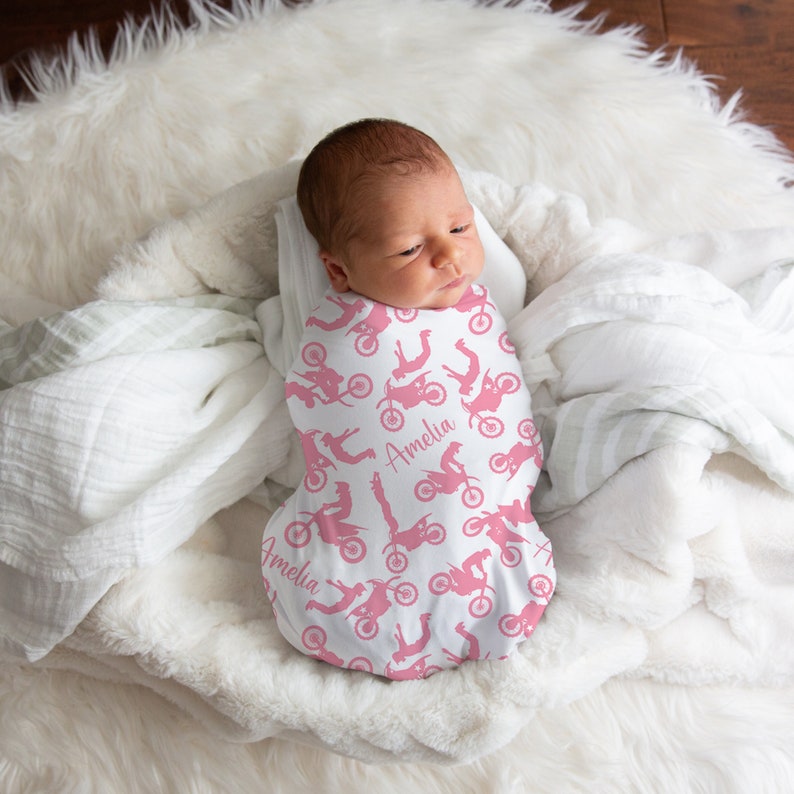 May include: A white swaddle with a pink and white motocross design. The swaddle has the name "Amelia" printed on it. A baby is sleeping inside the swaddle.