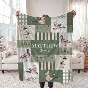 May include: A personalized blanket featuring a patchwork design with green and white squares and rectangles. The blanket showcases watercolor illustrations of ducks, foliage, and the name "MATTHEW JAMES" in white lettering. The blanket is held up by a person.