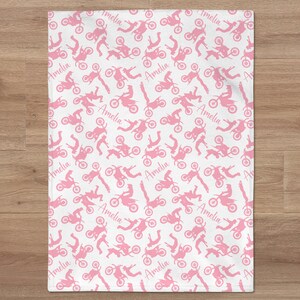 May include: Pink and white baby blanket with a repeating pattern of pink silhouettes of motocross riders and the name "Amelia" throughout.
