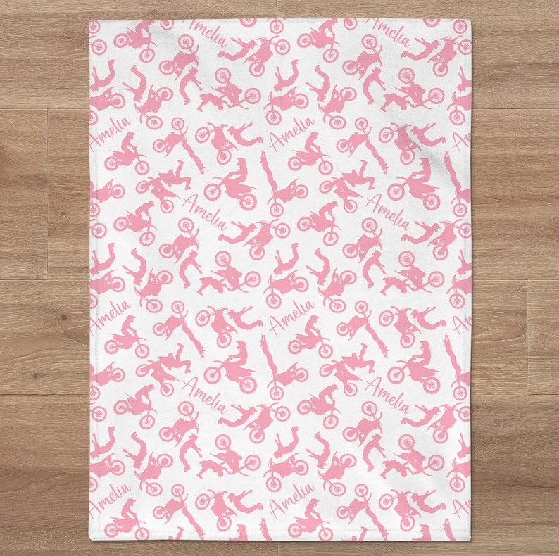 May include: Pink and white baby blanket with a repeating pattern of pink silhouettes of motocross riders and the name "Amelia" throughout.