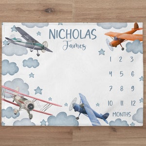 May include: A white baby milestone blanket with blue and white clouds, stars, and airplanes. The blanket has a blue border and the text "NICHOLAS James" and numbers 1-12 for each month.