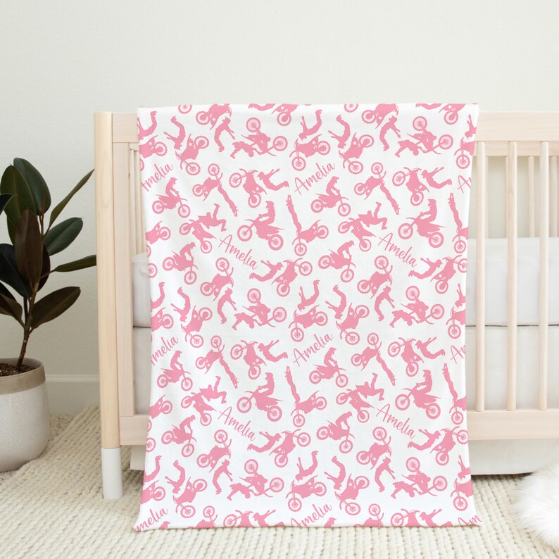 May include: A white baby blanket with a pink and white pattern of motocross riders. The blanket is personalized with the name "Amelia" repeated throughout the pattern.
