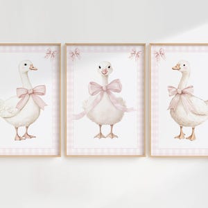 K&ouml;nnte beinhalten: Set aus drei gerahmten Kunstdrucken mit Aquarellillustrationen von wei&szlig;en G&auml;nsen mit rosa Schleifen. Jede Gans ist vor einem rosa Vichy-Hintergrund platziert und in hellem Holz gerahmt. Dekorative Schleifen befinden sich oben an jedem Rahmen.