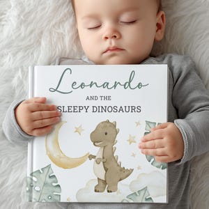 May include: A children's book titled "Leonardo and the Sleepy Dinosaurs" features a watercolor illustration of a friendly dinosaur, a crescent moon, stars, and clouds. The book's cover has a white background with the title in a script font.