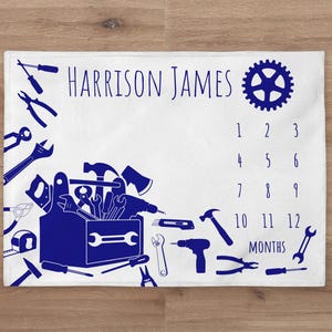 May include: White baby milestone blanket with a blue toolbox design and the text "HARRISON JAMES" at the top. The blanket has a number chart for each month of the year, with a gear design in the top right corner.