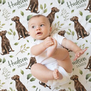 May include: A baby in a white onesie lies on a white sheet patterned with brown Labrador retrievers, green leaves, and the name "Hudson JAMES" in script. The baby is looking up with one foot in the air.