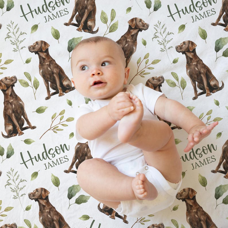 May include: A baby in a white onesie lies on a white sheet patterned with brown Labrador retrievers, green leaves, and the name "Hudson JAMES" in script. The baby is looking up with one foot in the air.
