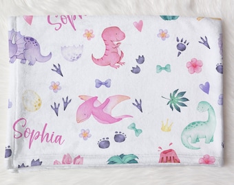 May include: A white baby blanket with a pink, green and purple dinosaur pattern. The blanket has the name "Sophia" written on it twice.
