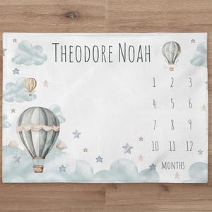 May include: A white baby milestone blanket with a blue and white hot air balloon design. The blanket features clouds, stars, and the text "Theodore Noah" and "Months". Numbers 1 through 12 are printed on the blanket.