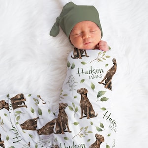 May include: A newborn baby wrapped in a white swaddle blanket and wearing a green cap. The blanket features a watercolor design of brown dogs, green leaves, and the name "Hudson James".
