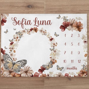 May include: A white baby milestone blanket with a floral wreath and butterflies. The blanket has the name "Sofia Luna" and numbers 1-12 for each month.