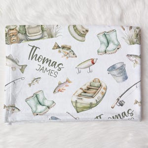 May include: White baby blanket with a fishing theme. Features illustrations of fish, fishing gear, a small boat, and the name "Thomas JAMES" in green script. Includes a fishing hat, boots, and fishing lures.