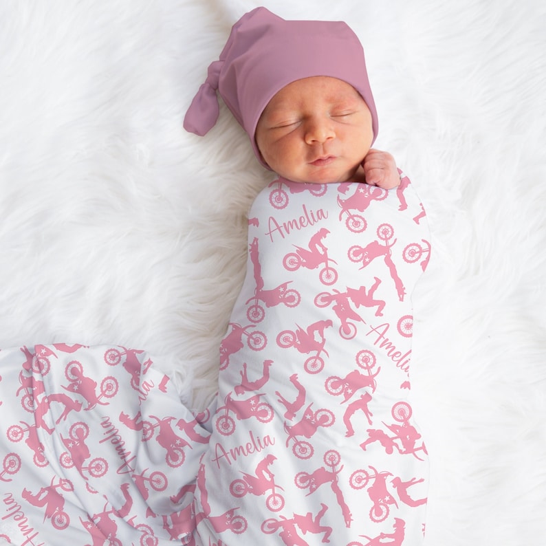 May include: A pink and white baby swaddle blanket with a pink motocross rider pattern and the name "Amelia" repeated throughout. A pink knotted hat is on the baby's head.