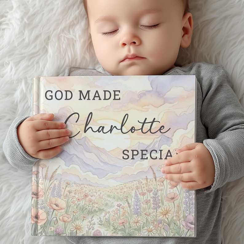 May include: A baby rests while holding a book titled "GOD MADE Charlotte SPECIAL." The book cover features a watercolor landscape with mountains, flowers, and a sunset. The baby is wearing a gray long-sleeved shirt.
