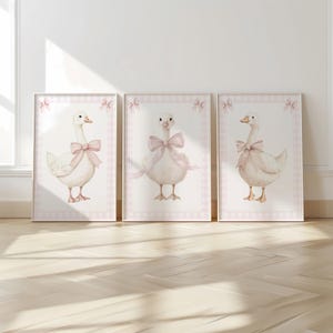 K&ouml;nnte beinhalten: Drei gerahmte Aquarelldrucke mit bezaubernden wei&szlig;en G&auml;nsen, die mit rosa Schleifen geschm&uuml;ckt sind. Jeder Druck hat einen rosa Vichy-Karo-Rand mit dekorativen Schleifen. Das Kunstwerk wird auf einem hellen Holzboden ausgestellt.