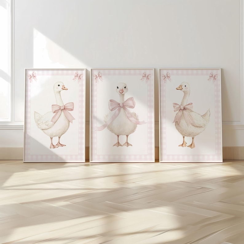 K&ouml;nnte beinhalten: Drei gerahmte Aquarelldrucke mit bezaubernden wei&szlig;en G&auml;nsen, die mit rosa Schleifen geschm&uuml;ckt sind. Jeder Druck hat einen rosa Vichy-Karo-Rand mit dekorativen Schleifen. Das Kunstwerk wird auf einem hellen Holzboden ausgestellt.